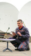 © wedmoments.stock - Technician in gray coveralls kneeling on rooftop while using tablet to install satellite internet dish, large satellite dishes visible in the background