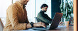 © Prostock-studio - Two men are seated at desks in a modern office. One man is focused on his laptop, while the other sits in the background. The scene shows a typical work environment with natural light.