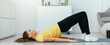 © Prostock-studio - A woman lies on a mat in her living room while performing the bridge exercise. She keeps her feet flat and lifts her hips off the ground. Sunlight shines through a window.