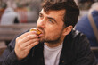 © zvkate - Close up of hungry middle-aged man with blue eyes and thoughtful facial expression eating delicious cookie or pastry outdoors, handsome male in black jacket enjoying sweet snack at street cafe