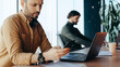 © Prostock-studio - Two men are seated at desks in an office setting. One man looks at his smartphone, while the other works on a laptop. Sunlight streams through the windows, creating an urban workspace.