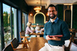 © djile - Smiling young adult man using digital tablet while standing in stylish cafe with warm interior design.