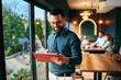 © djile - Young man browsing online content on tablet while standing near window in contemporary cafe setting.