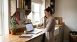 © Alina Sukach - A woman speaking on her phone in a cozy kitchen workspace, open laptop on the counter, warm natural light, soft wood textures and everyday home details creating a relaxed productive atmosphere,