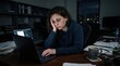© Cealv - An exhausted woman works late on her laptop at a messy desk in a dark office with city lights.