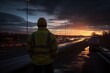 © juliars - Worker in safety vest and hard hat standing beside a highway at dusk with long exposure light trails