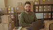 © Krakenimages.com - Hispanic man with beard smiles, hands clasped on laptop amid stacked shipping boxes and scanner in a building; entrepreneurial confidence.
