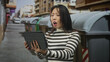 © Krakenimages.com - Woman holding tablet with one hand while the other covers her mouth, striped sweater, standing on a narrow urban street next to metal trash bins and parked cars; shock vulnerability.