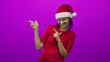 © Krakenimages.com - Woman pointing finger guns with both hands, wearing a santa hat and red shirt against a vibrant purple studio backdrop; playful cheer.