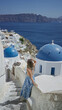 © Krakenimages.com - Young hispanic brunette woman smiling and holding a railing on a church building in santorini, overlooking blue domes and the sea caldera; serenity travel escape.
