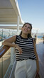 © Krakenimages.com - Woman leaning on ship railing, smiling and looking up on a cruise terrace wearing striped top and white pants, sunglasses on head and holding a bag; carefree joy.