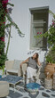 © Krakenimages.com - Woman seated on a wicker chair touching her hair on a white building terrace in mykonos, overlooking stone patio and potted plants; calm leisure.