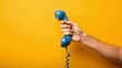 © olga_demina - A close-up of a hand grasping a blue telephone receiver with a coiled cord, set against a solid yellow backdrop, evoking a sense of nostalgia and simplicity in communication technology.