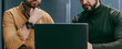 © Prostock-studio - Two men are sitting close together at a table. They are looking at a laptop screen and discussing something important. The setting is an office with a modern design.