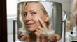 © Frame of the Dark - Beautiful mature woman applying anti-aging face cream in mirror, senior skincare beauty routine