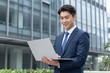 © Studicon - Young asian businessman smiles while holding a laptop outdoors in front of a modern office building