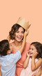 © Milan - Vertical portrait of a happy mother being crowned by her children. Smiling woman with son and daughter on peach background. Queen for a day Mother's Day concept