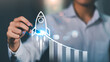 © chaylek - A person draws a rocket launching upward alongside a rising bar chart, symbolizing startup growth, business acceleration, performance metrics, innovation strategy, exponential success driven by data