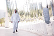 © .shock - Smiling Arab man in traditional dress walking by reflective modern architecture in an urban park