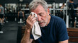 © Cliff - Portrait of a tired but satisfied senior man wiping his face with a towel after an intense workout. He sits on a bench in the gym, his skin covered in sweat, his face showing fatigue and joy