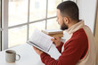 © Pixel-Shot - Handsome young man with cup of coffee reading book at table near window