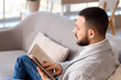 © Pixel-Shot - Handsome young man reading book on grey sofa at home