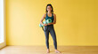 © Cliff - Portrait of a joyful young woman in stylish sportswear holding a rolled up yoga mat, bright yellow background, studio shot, concept of healthy lifestyle, fitness motivation and positive energy