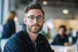 © Fox - Confident young man with glasses and beard sitting in modern office environment, looking at camera. Professional millennial in casual denim jacket at creative workplace