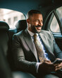 © Kowit - A well-dressed man in a suit and tie smiles while sitting in the backseat of a car, holding a smartphone and looking out the window.