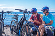 © luciano - Happy senior couple with helmets sitting on a rocky beach before riding with their electric bicycles. The man looking at smartphone, the lady eating an apple. Authentic elderly retired life concept.