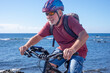 © luciano - Active senior man cycling along a scenic seaside coastline on a sunny day. Wearing a helmet, sunglasses, and casual sportswear, enjoying a healthy outdoor lifestyle and the freedom