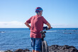 © luciano - Back view of active senior man cycling along a scenic seaside coastline on a sunny day. Wearing a helmet,  and casual sportswear, enjoying a healthy outdoor lifestyle and the freedom