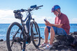 © luciano - Smiling active senior man with bicycle sitting resting along a scenic seaside coastline using phone. Wearing a helmet and casual sportswear, enjoying a healthy outdoor lifestyle and the freedom