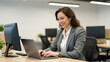 © NAD SANG PAP - Smiling businesswoman in gray suit working on laptop at modern office desk with desktop monitor and potted plant, showing positive and focused expression