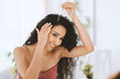 © Prostock-studio - A woman is using a dropper to apply hair oil to her scalp while looking in the mirror. She has curly hair and a bright smile. The setting is light and filled with plants.