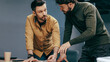 © Prostock-studio - Two men are engaged in a discussion at a table in an office. They are looking at documents and sharing ideas about a project. The setting is a workspace with a relaxed atmosphere.