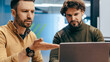 © Prostock-studio - Two men are seated at a table in an office. One man gestures as he speaks, while the other focuses on the laptop screen. They are engaged in a conversation about their work.