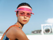© SHOTPRIME STUDIO - Portrait of a youthful woman on a sunny beach wearing a pink visor and colorful athletic top. Close framing highlights smooth skin, confident gaze, and summer mood.