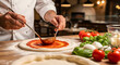 © Aura - Close up of a chef in white uniform using a ladle to spread red tomato sauce in a circular motion on raw pizza base
