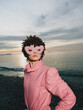 © SHOTPRIME STUDIO - portrait on a beach at sunset featuring a person in a pink jacket wearing a playful mask, a bold fashion moment by the sea under warm light and cinematic mood