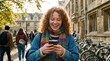 © Влада Яковенко - A smiling teenage girl with curly ginger hair using a smartphone outdoors in vertical composition, casual student style, campus surroundings and fresh daylight, playful mobile-gaming mood with
