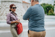 © qunica.com - A professional woman speaks with a colleague by a decorative fountain during a business conversation. They chat outdoors while she holds a red bag and a smartphone.