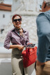 © qunica.com - A smiling businesswoman holds a tablet and red briefcase while speaking to a colleague. Casual outdoor business conversation captured near a fountain.
