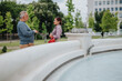 © qunica.com - A middle-aged man and woman have a business conversation standing beside a city fountain. The colleagues converse casually in a park setting near decorative water features.