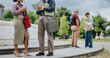 © qunica.com - Two business colleagues stand talking while holding notebooks and bags. A casual outdoor work meeting unfolds on park steps with coworkers nearby.