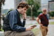 © qunica.com - A business man smokes a cigarette while checking a tablet, focused on the screen. He sits on a bench in an urban setting with a blurred passerby in the background.