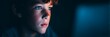 © Tanakrit - A close-up of a focused young boy with freckles, illuminated by blue light, gazing intently at a screen in a dark environment.
