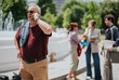 © qunica.com - A mature businessman stands by a city fountain speaking on his smartphone. In the background colleagues converse and review documents during a casual outdoor business meeting.