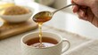 © nunoi - A hand drizzling honey from a spoon into a cup of tea, with a bowl of brown sugar in the background on a light cloth surface.