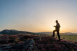 © antasfoto - silhouette of a man photographing in the mountains, A person is silhouetted against the setting sun while walking along a rocky, flower-covered ridge overlooking a vast landscape. Sardinia Italy